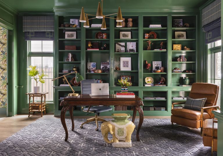 Home office with green walls and shelving, grey rug, antique desk, leather armchair and pendant light
