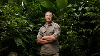 Paddy McGuinness wearing khaki coloured clothing with his arms folded smiling at the camera in a green jungle.