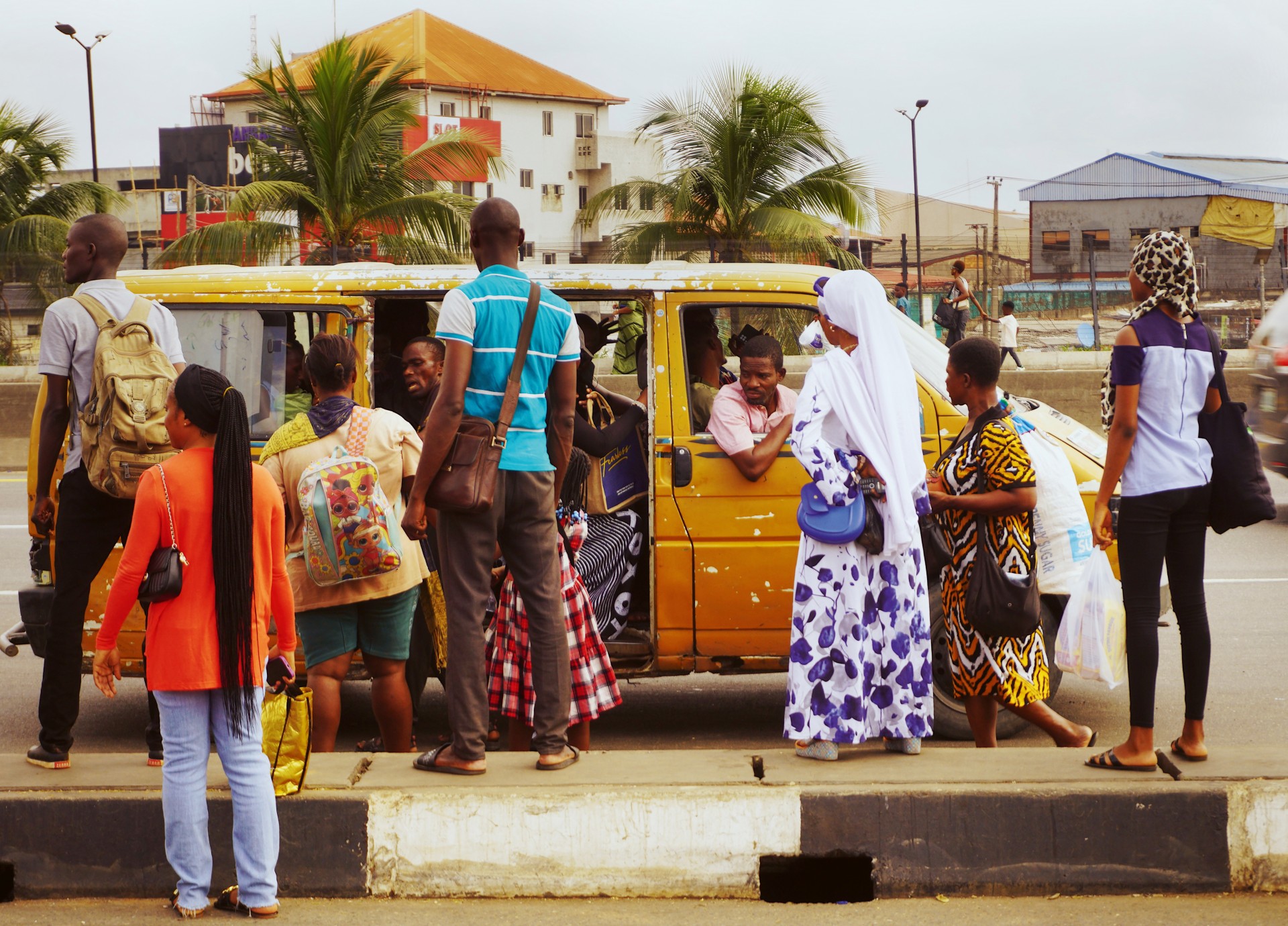 A line of commuters waits at a roadside bus stop beside a battered yellow minibus, most of them facing away from the camera.