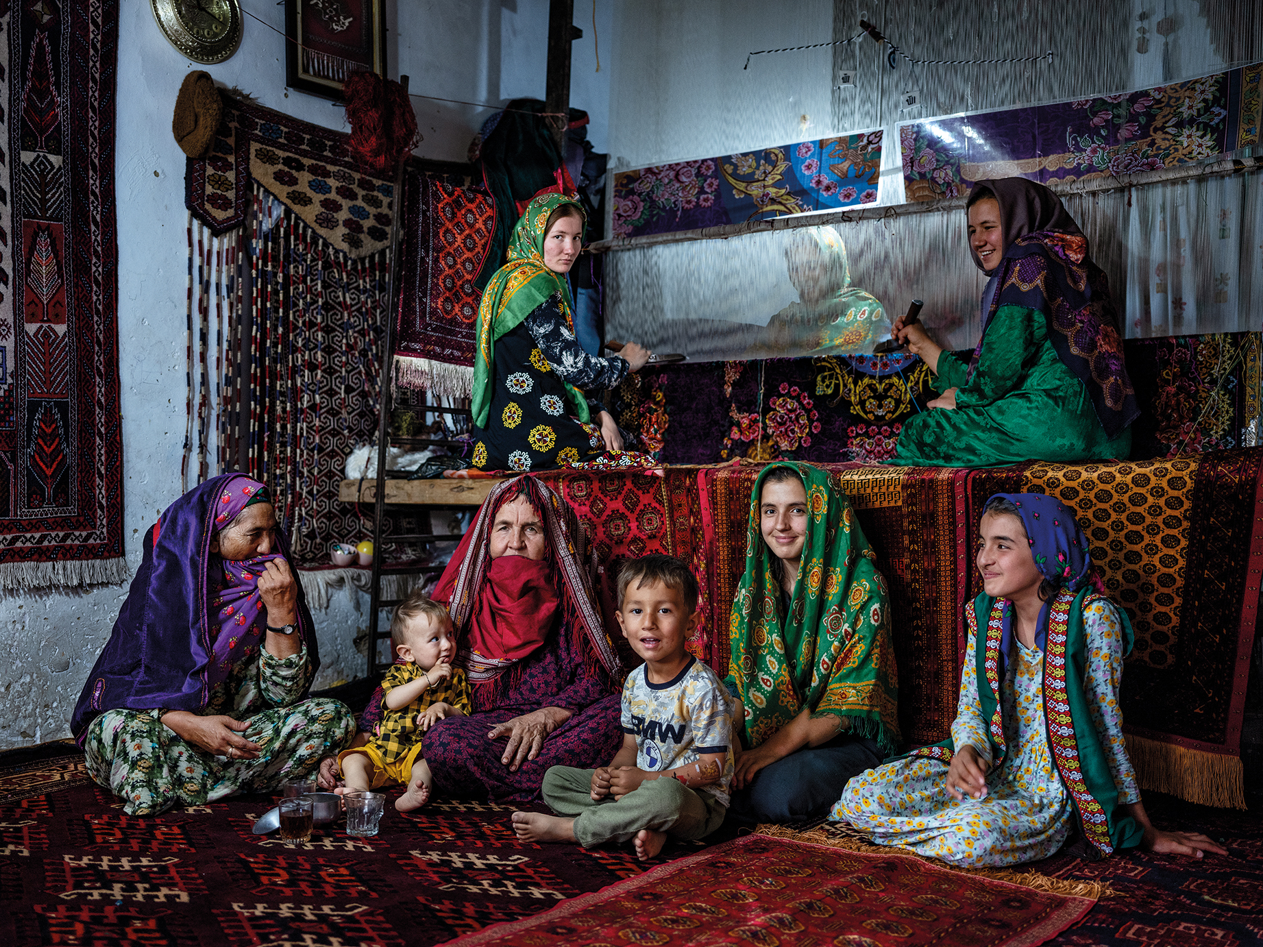 A group of women and children dressed in vibrant traditional clothing, seated on intricately patterned carpets in a warmly lit room