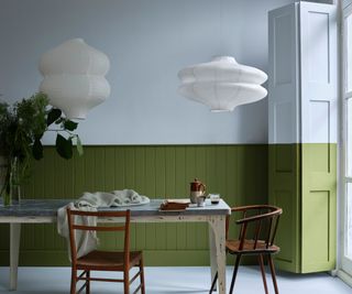 dining space with light blue upper half of walls and dark green panelling on the lower. dining table with two vintage wooden chairs and two white paper pendant lights