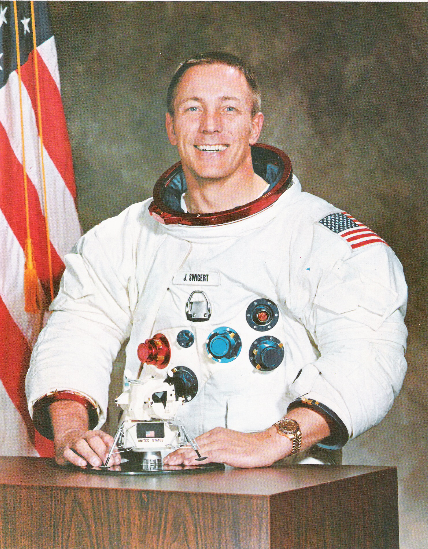 jack swigert standing in a spacesuit and smiling. he holds a model lunar rover in his hands, which is resting on a table