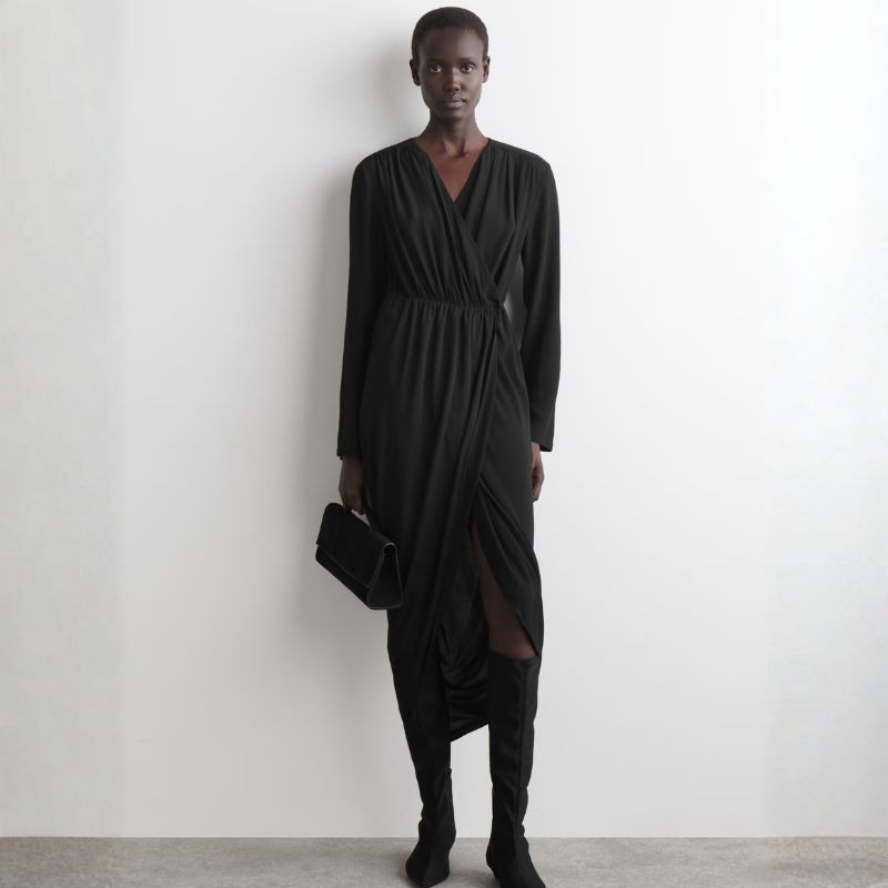 A woman stands against a wall wearing a COS dress and carrying a black handbag