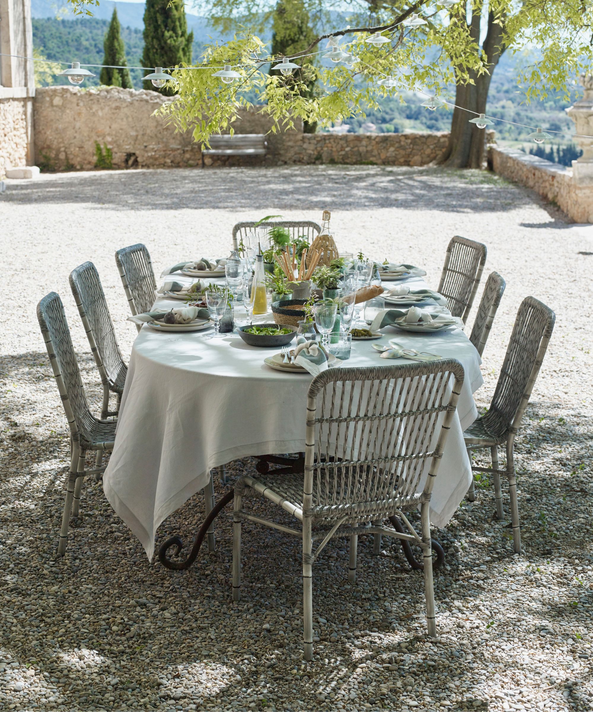 An outdoor dining set-up in a Mediterranean garden with trees, a white tablecloth over it, metal vintage chairs, a tablescape of green and white plates, glassware, and fresh green flowers.