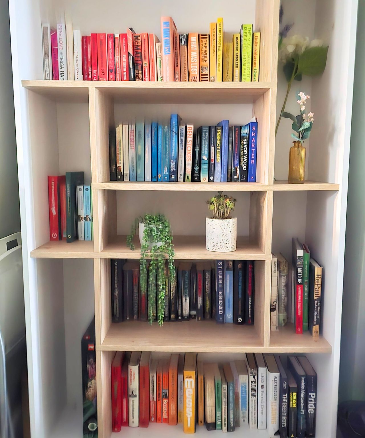 A white bookshelf with colorful books in rainbow order on it and faux plants in the middle of it