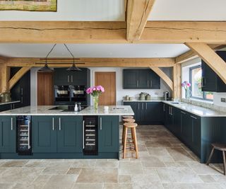 A new kitchen in an oak frame home with stone flooring and a large kitchen island. The cabinets are a dark teal and the worktops are light