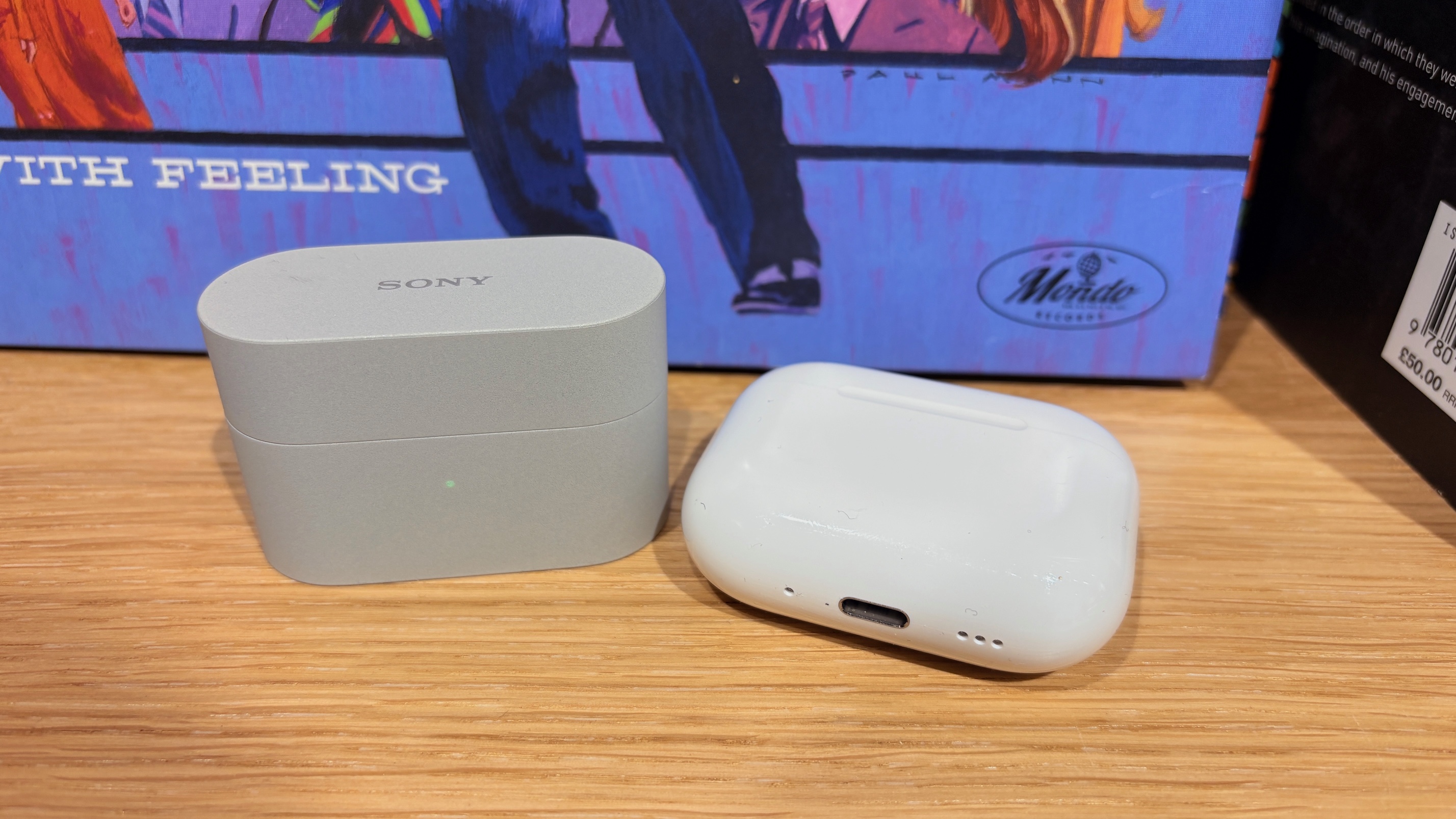 The charging cases of the Sony WF-1000XM6 and Apple AirPods Pro 3 next to each other on a wooden surface.