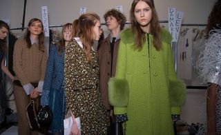 Female model in a green duffel coat with green fur cuffs and collar. Other models in the background