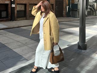 Marina wears white top and pants and black Mary Jane flats with a tan chore jacket and brown handbag.