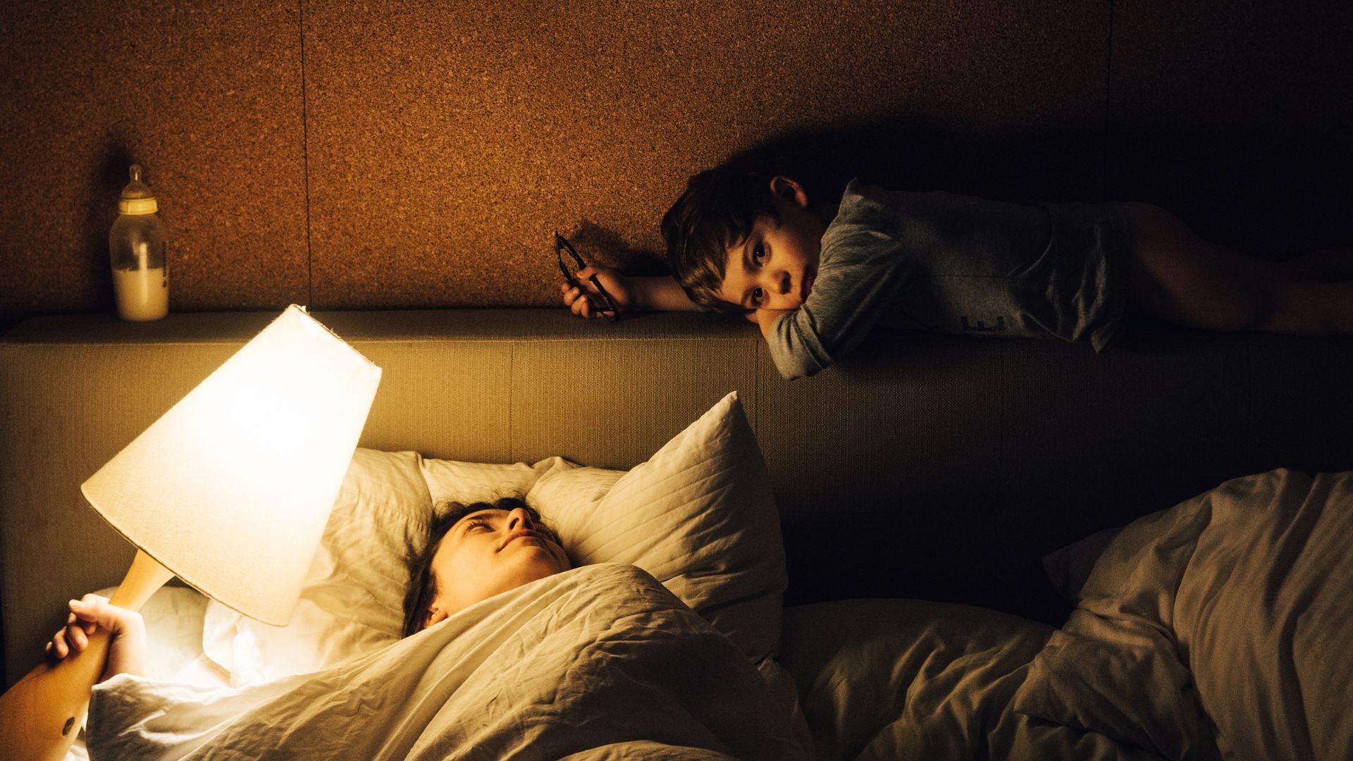 A young child rests on the headboard of their parents' bed in the middle of the night as the father looks up at them, holding a lamp to illuminate the child.