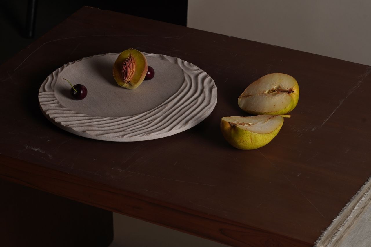 A white plate with a rough, organic finish contains half a peach and two grapes while sitting on a brown, wooden slab next to two half pears.