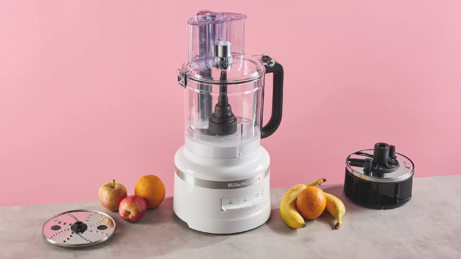 A white KitchenAid Classic 13 cup / 3.1l sitting on a stone effect surface and against a pink background, with the storage caddy containing a reversible slicing disc on the right side, and the reversible grating on the left.disc