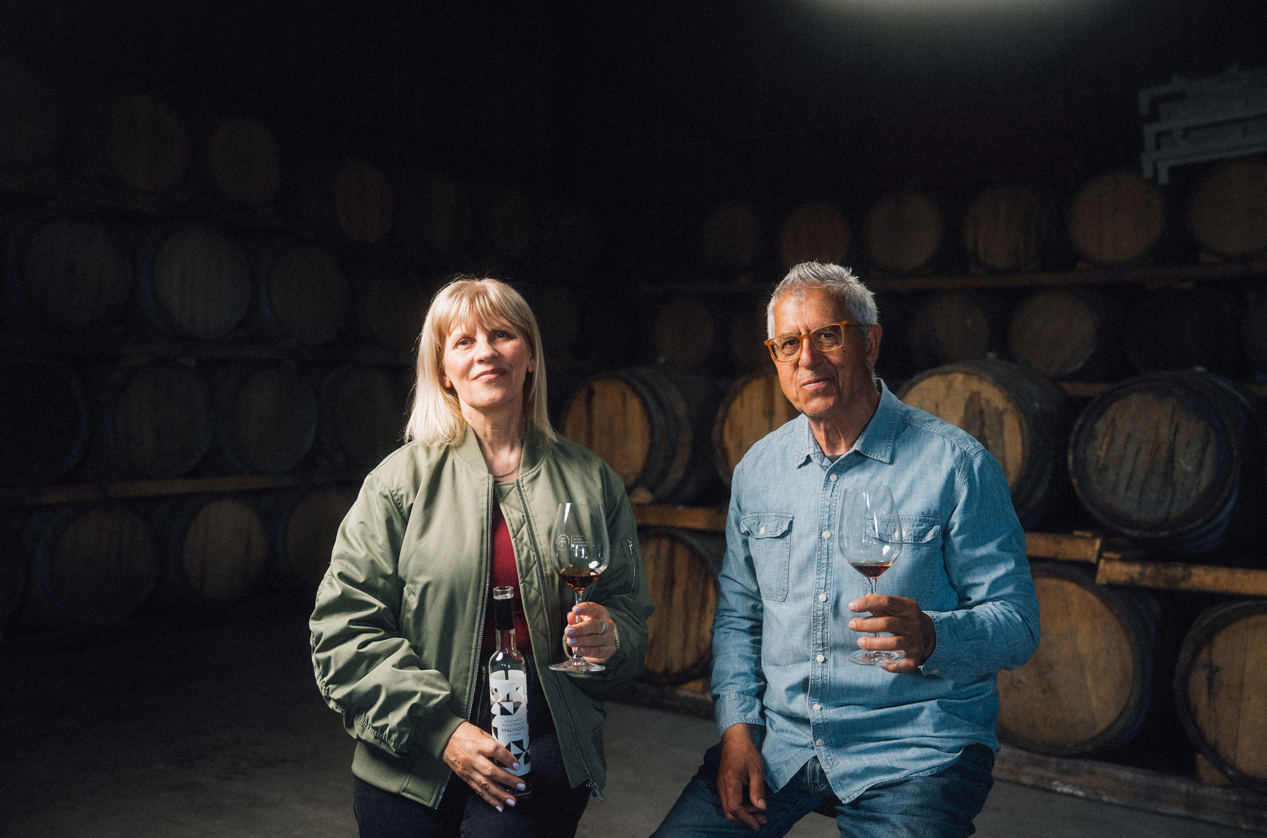 A couple holding wine glasses, sat in front of oak barrels