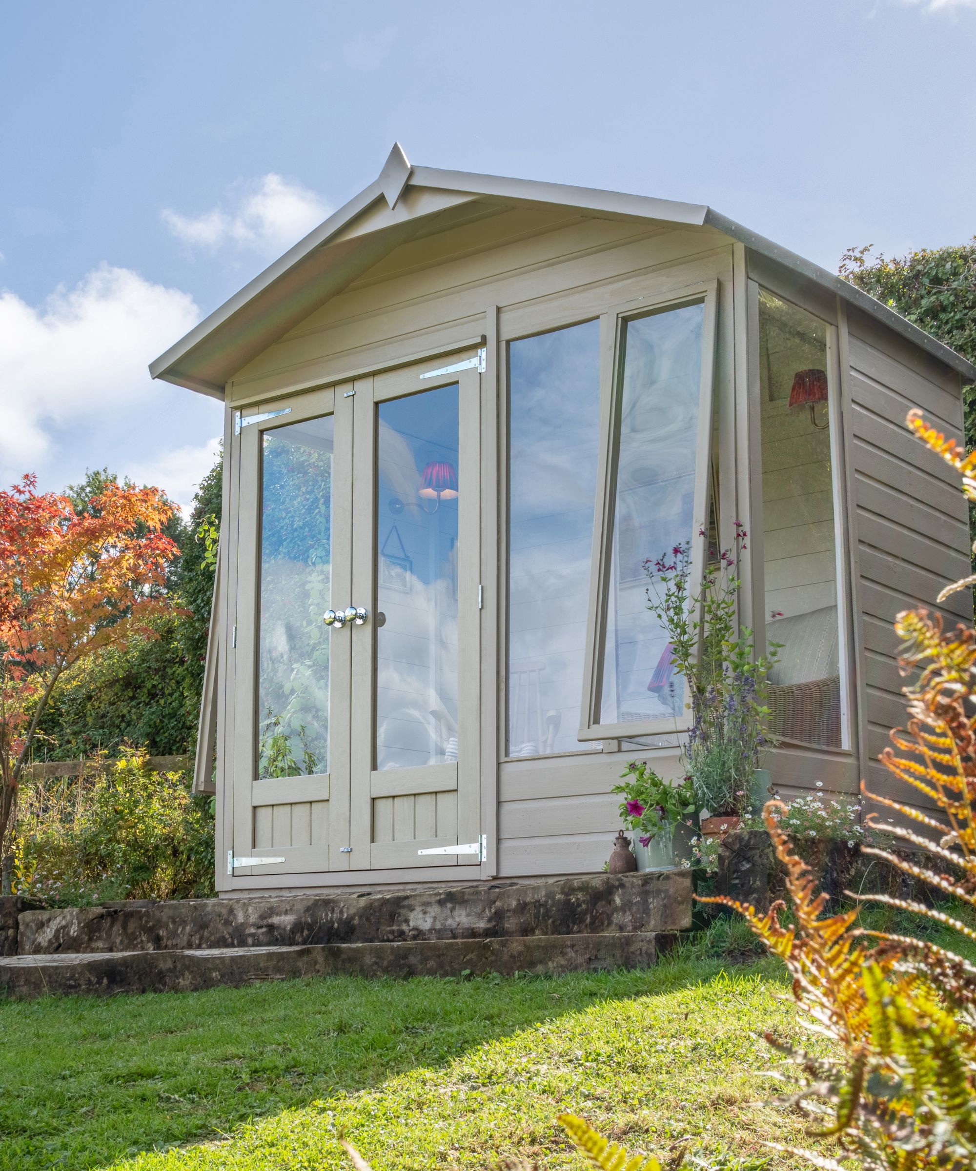 A sage green painted garden summerhouse with large glazed double doors and side windows, raised on a stone base and surrounded by autumnal planting and ferns.