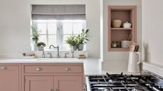 A pink and white kitchen with sink beneath window and gas burner stove