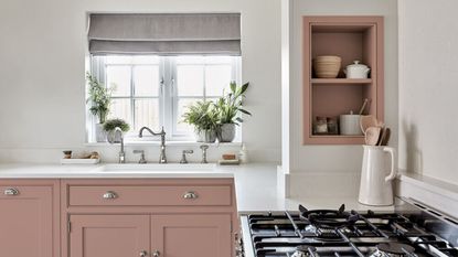 A pink and white kitchen with sink beneath window and gas burner stove