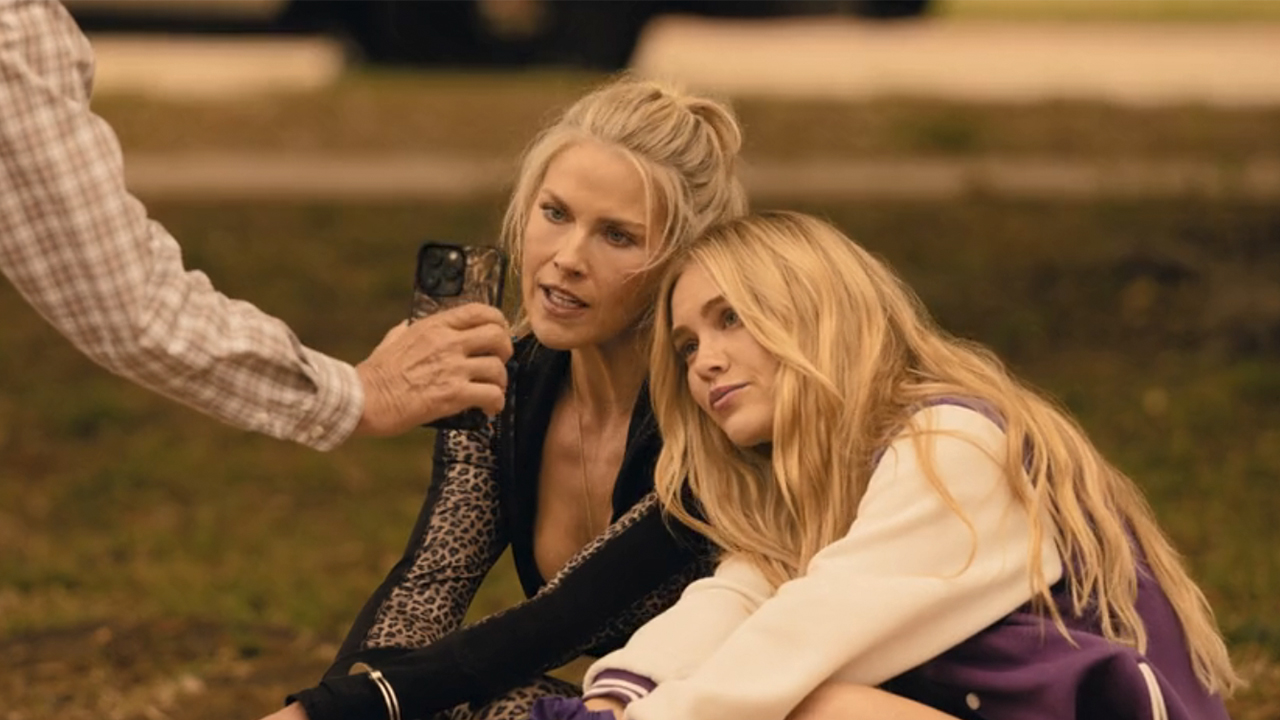 Michelle Randolph and Ali Larter talking into a phone while seated on the ground in Landman