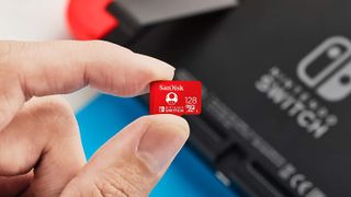 En hand håller upp ett rött Sandisk microSDXC över en Nintendo Switch-konsol i bakgrunden.