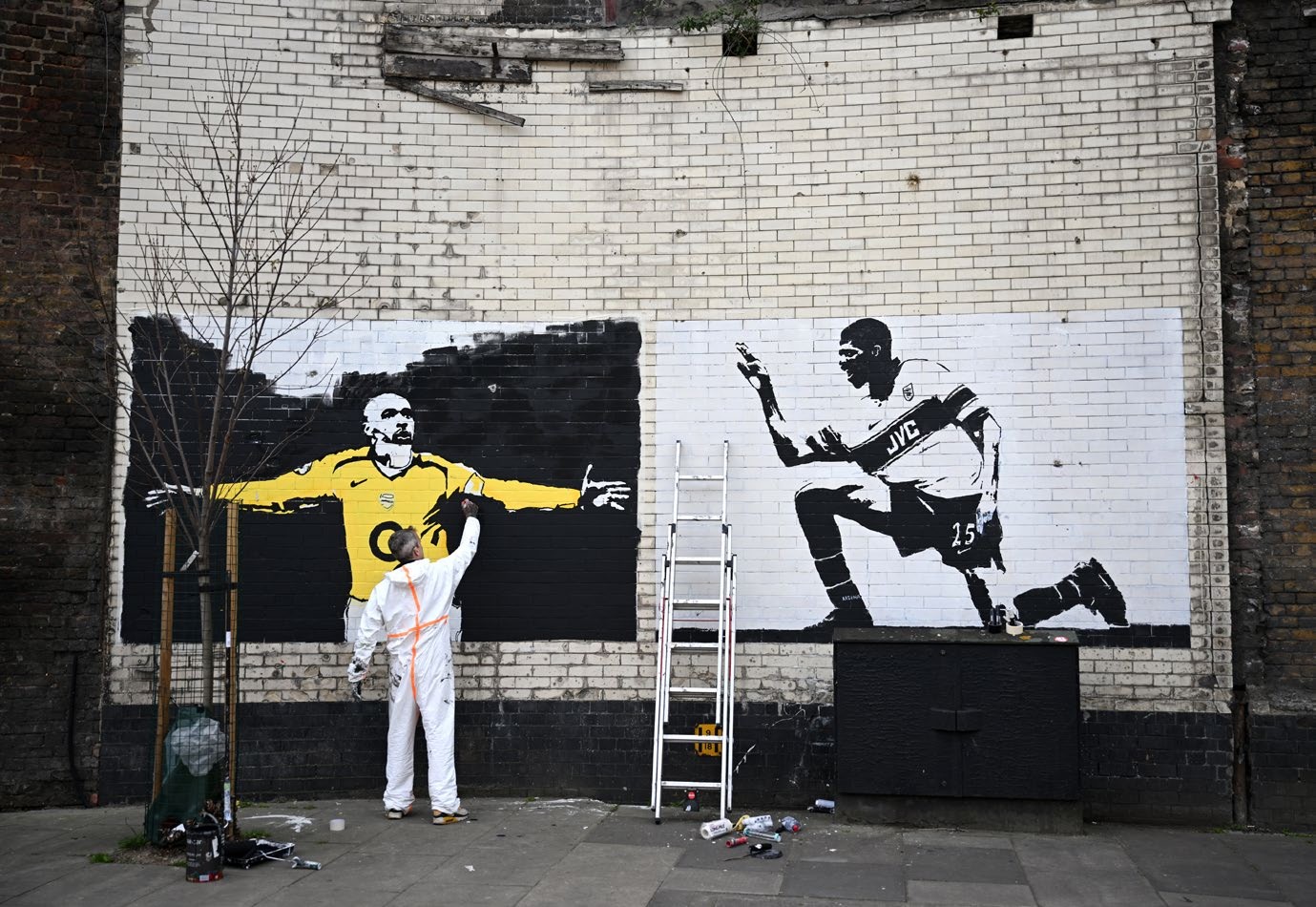 A street artist painting a mural depicting a goalkeeper in yellow and a football player in black and white on a brick wall, reflecting the vibrant street art culture inspired by local football legends.