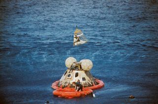 Sitting in a "Billy Pugh" net, Apollo 13 command module pilot Jack Swigert approaches the helicopter as his fellow astronaut Jim Lovell, the mission commander, waits below in the life raft. Above, lunar module pilot Fred Haise is already in the aircraft. Several U.S. Navy underwater demolition team swimmers aid in the recovery operations.
