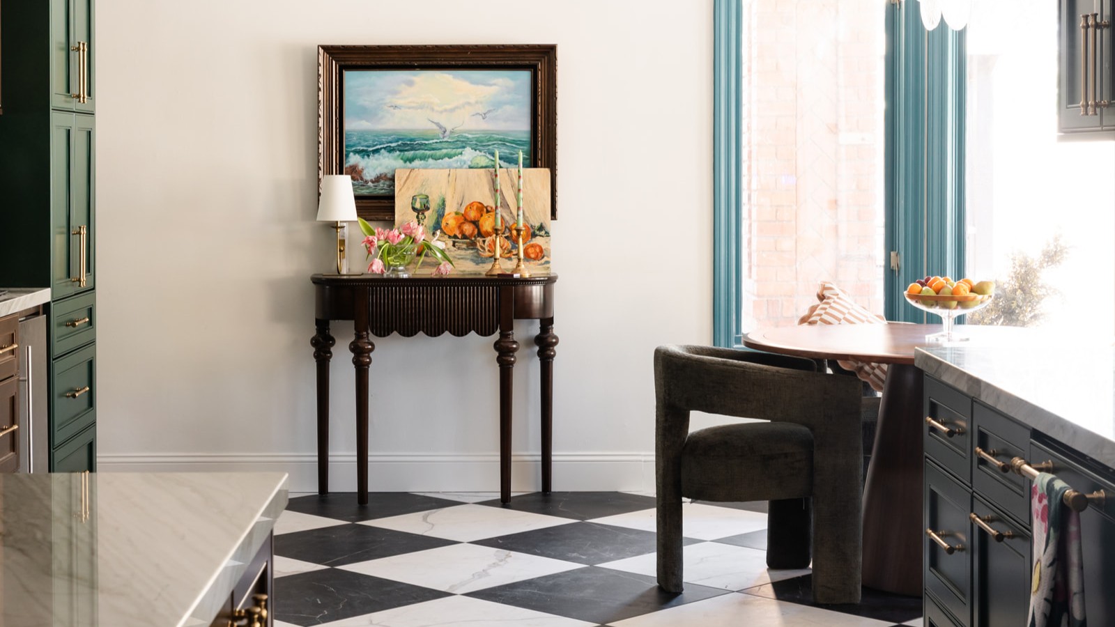 Kitchen/dining space with black-and-white tile floor. A side table is against the wall in the background with a lamp, flowers and candles, below a framed painting. Corners of shiny countertops are pictured in the foreground.