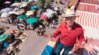 Phil Keoghan stands on a rooftop while explaining a challenge on The Amazing Race. He has on a straw hat and red button down and jeans.