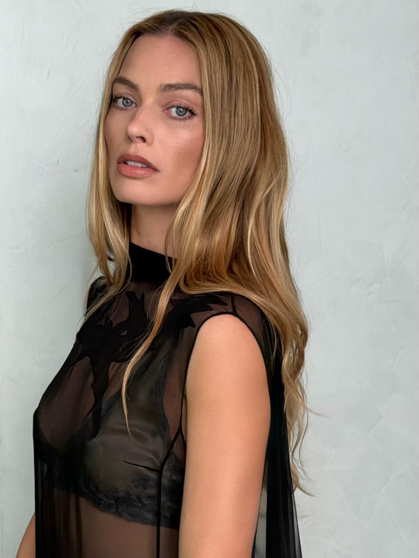 Margot Robbie with long, blonde, wavy hair wearing a black sheer dress