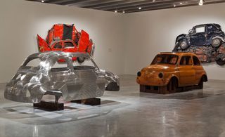 A silver and a yellow car body face each other, two other crushed cars hang on the walls