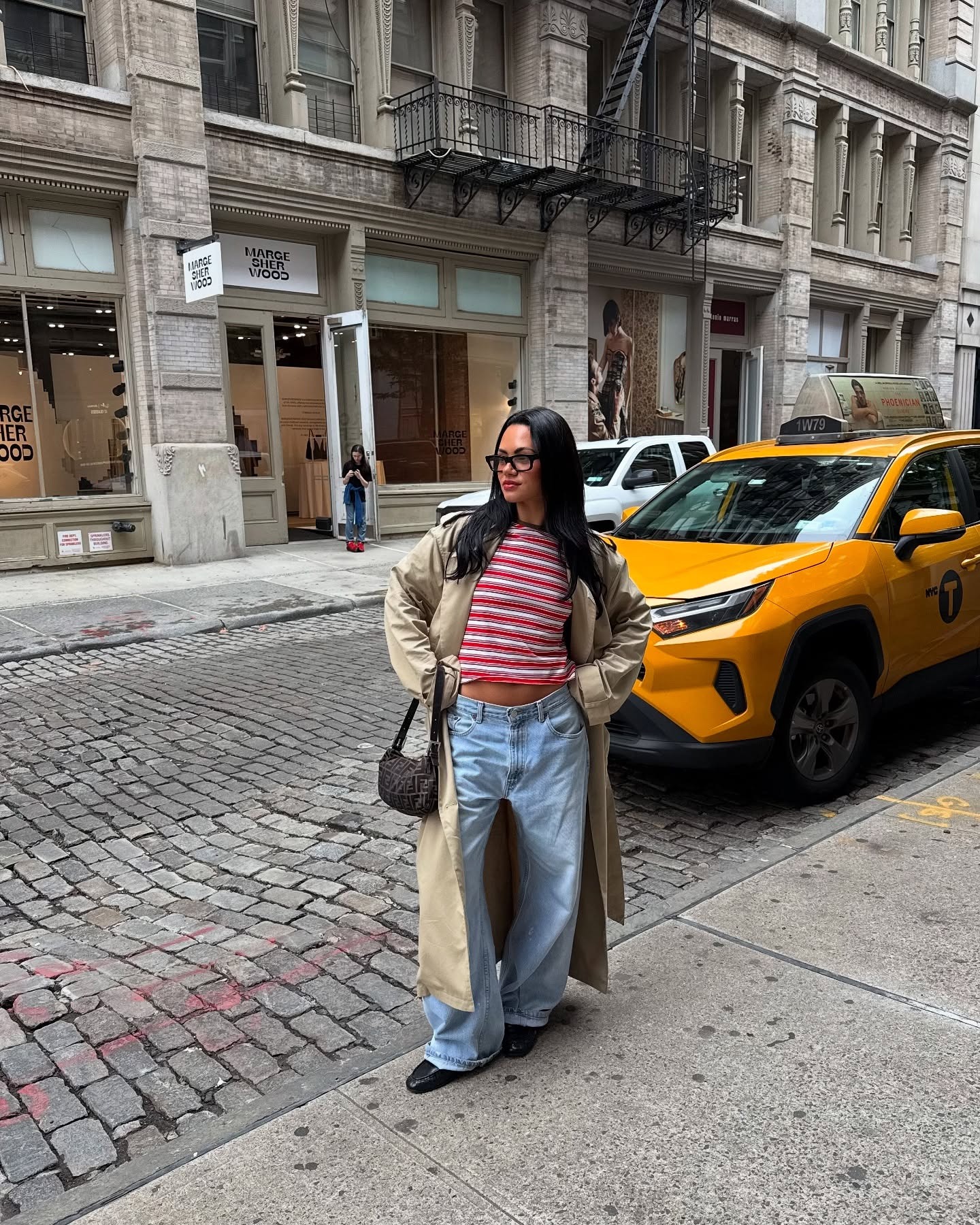 A woman wearing a striped shirt, a tan trench coat, and blue jeans in NYC.