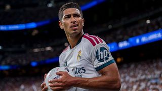Jude Bellingham, wearing the white of Real Madrid, carries a football and winks at the crowd after scoring a goal.