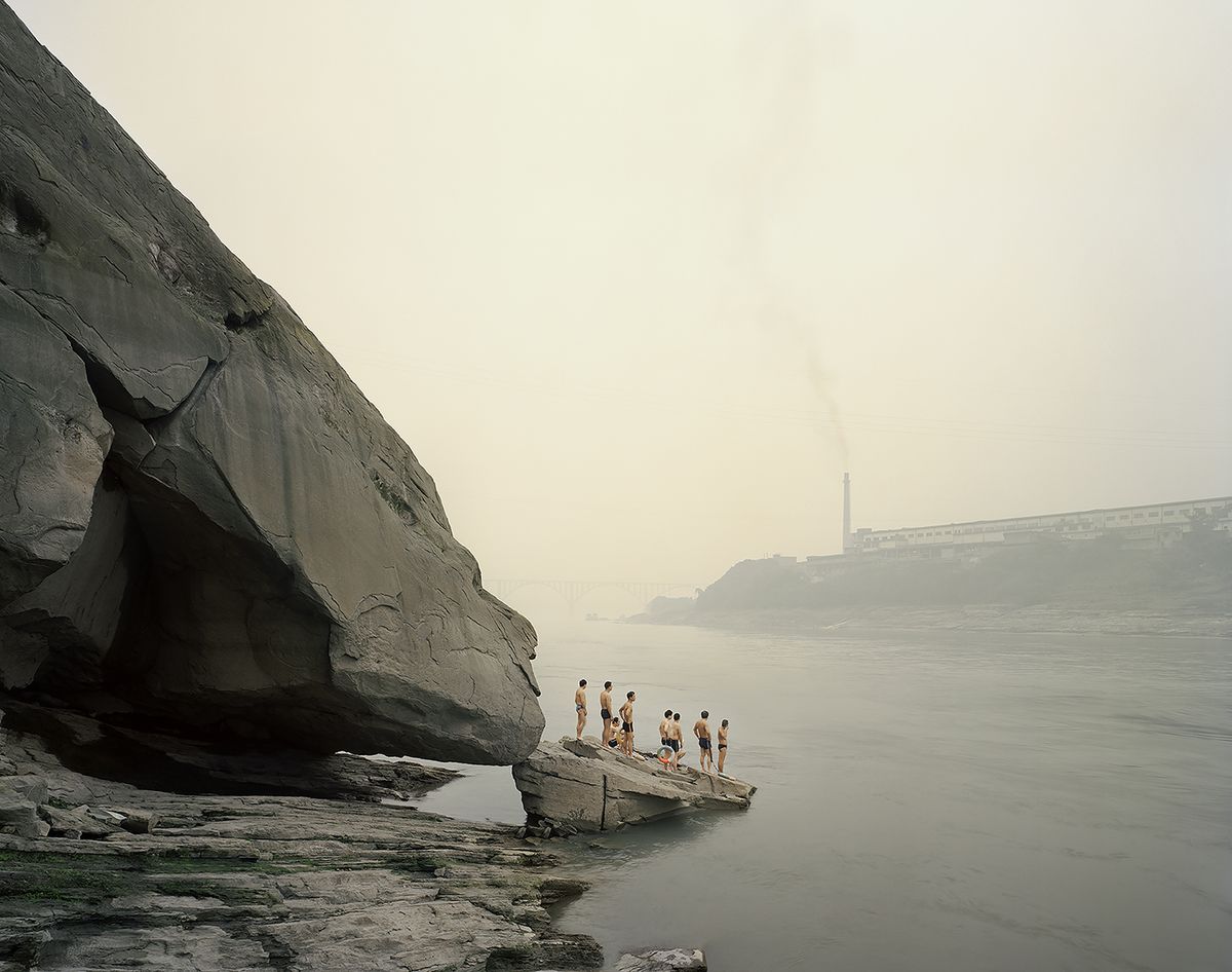 A small group of swimmers stands on a rock beside a huge overhanging boulder on a hazy river, with a smoking factory chimney and a bridge faint in the distance.