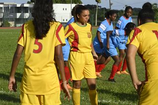 Grace Devon (Rita Bernard-Shaw) stands on a football pitch with the rest of the Saint Marie Women's Youth Team warming up. They are wearing a yellow kit with red stripes down the sides of their sleeves. The Saint Auguste team are visible in the background, in a sky-blue kit with white stripes on the sleeves.