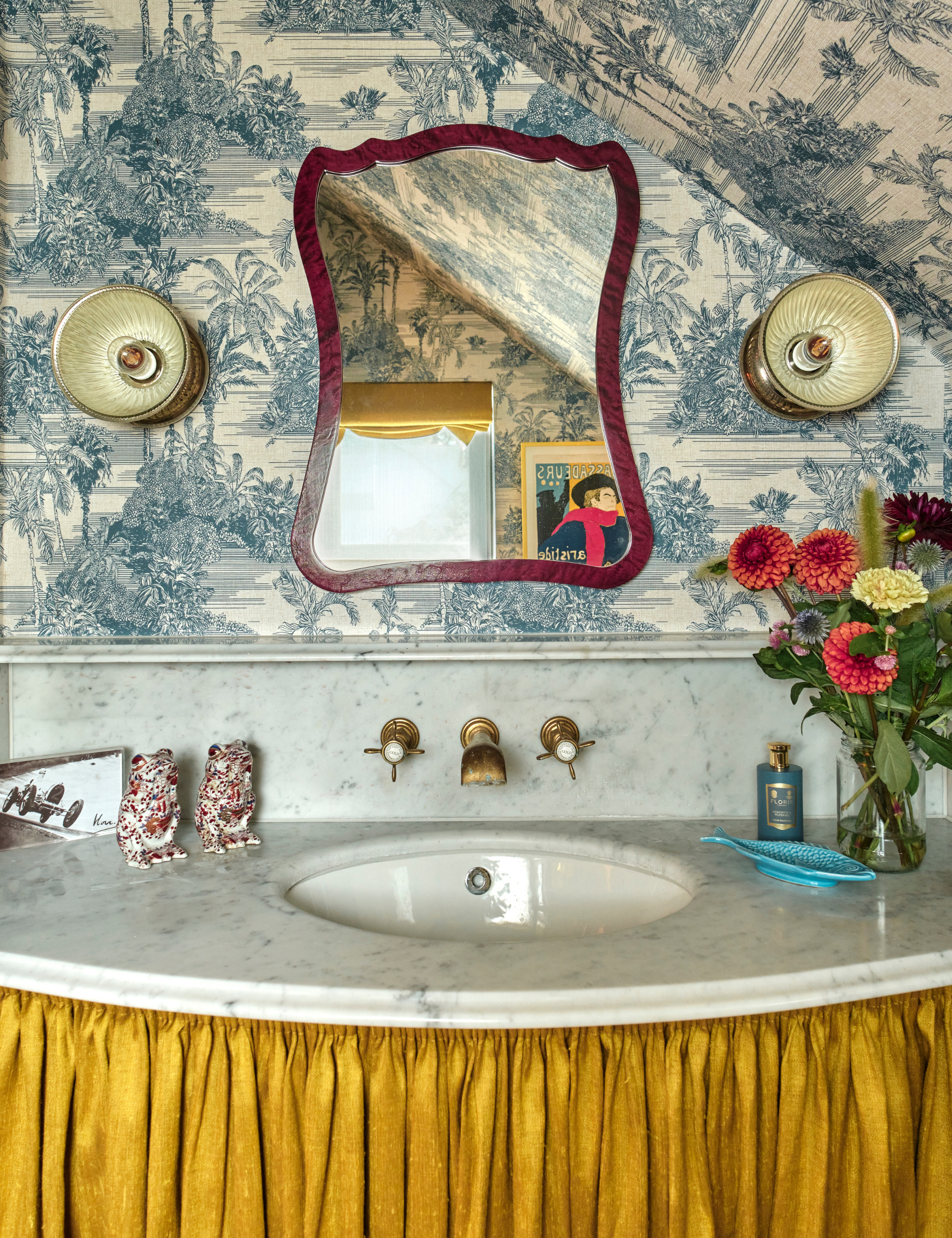 Bathroom sink vignette featuring a mustard yellow sink skirt, patterned toile wallpaper, sculptural vanity mirror, and a vase of fresh flowers.