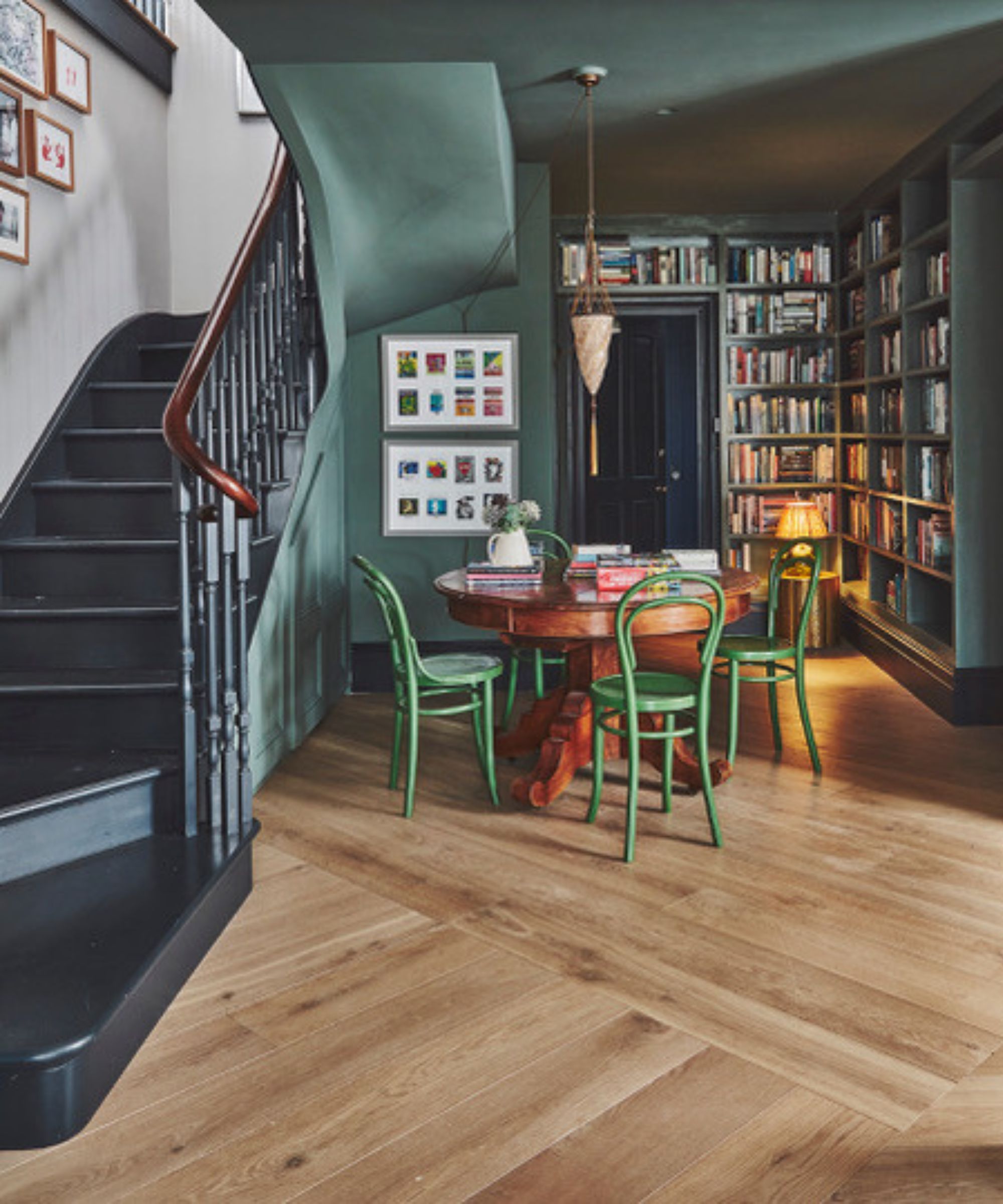 Cosy reading nook beneath a curved staircase, with dark green walls, floor-to-ceiling bookshelves and vibrant green café chairs adding character.