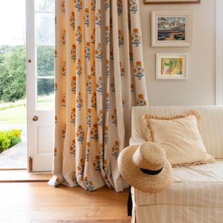 colour ideas for cottagecore decor, patterned cream curtains in cream living room, yellow stripe sofa, patterned yellow cushions, artwork, wooden floor, open door to garden