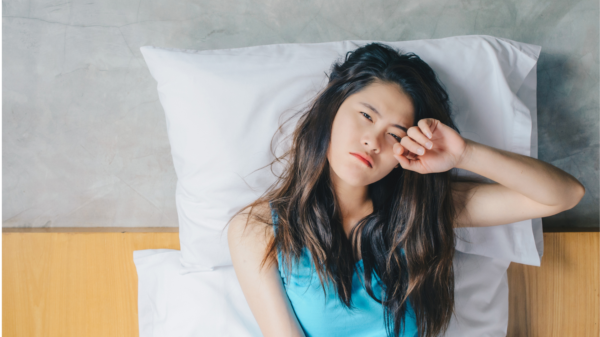 A woman sits up in bed looking tired and unhappy, she has two pillows supporting her as she sits up.