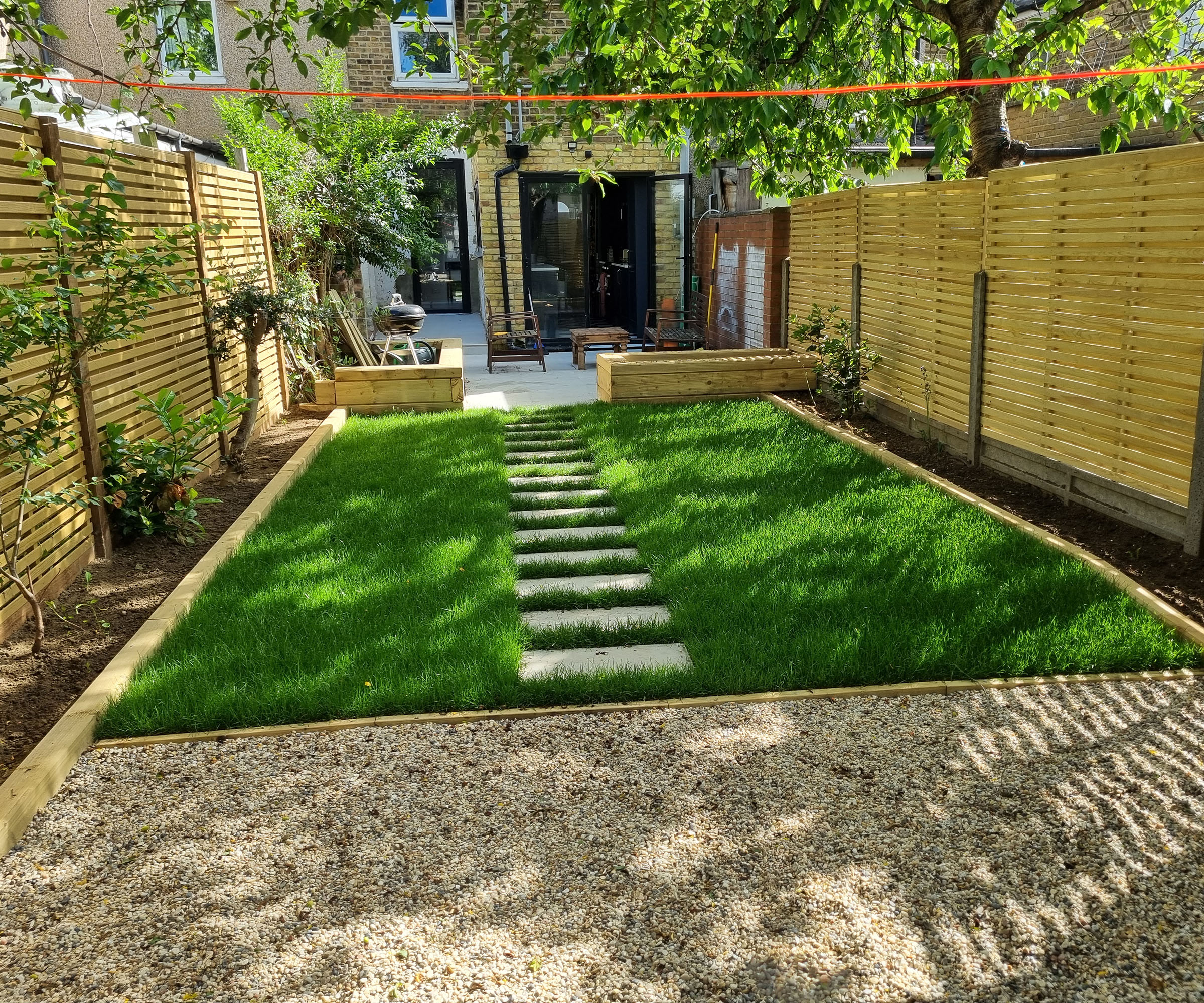 A landscape shot of a new garden makeover looking back towards a house with a gravel area, turf with stepping stone path and garden beds either side