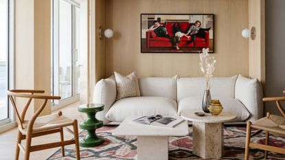 A naturally lit living room with a softly shaped white lounge sofa, a red, blue, and cream rug, wooden chairs, two marble coffee tables, one round, one squared, and spherical scones.