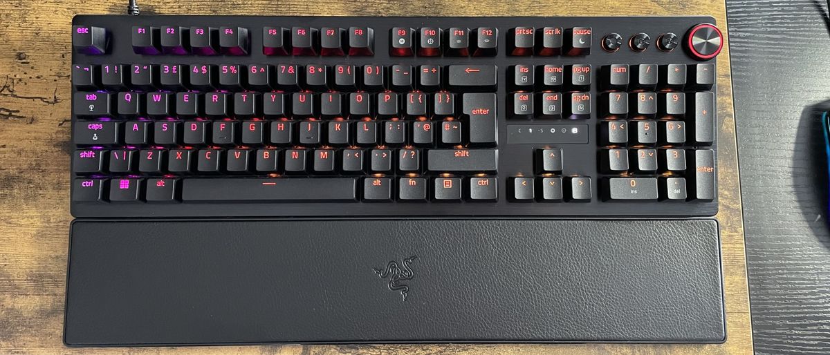 The Razer Huntsman V3 Pro shown on a wood effect desk top with purple and orange backlighting behind the keys.