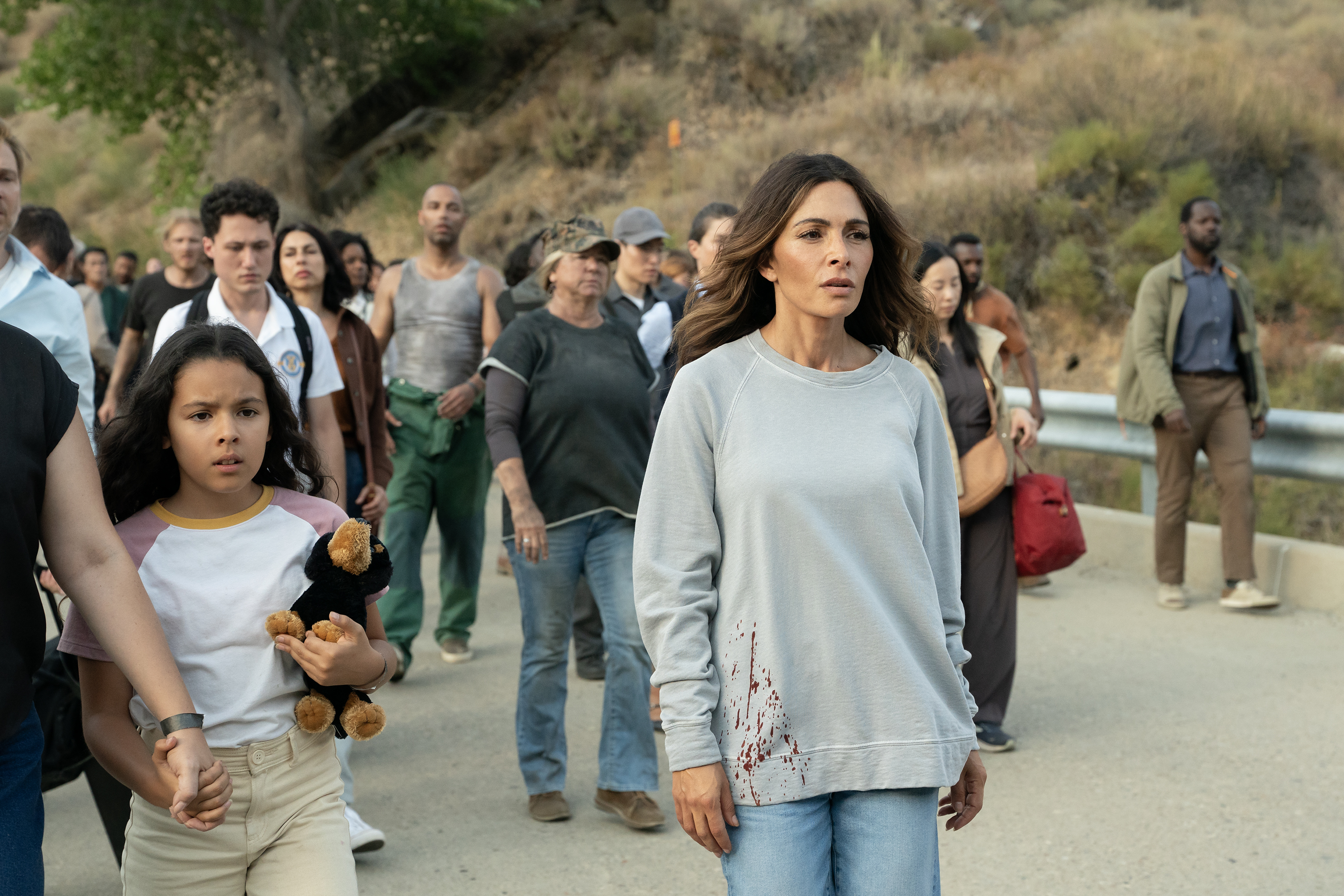 A woman wearing a bloody sweatshirt (Sarah Shahi as Dr. Torabi) looks dazes while walking down a highway with dozens of people. A young girl next to her holds a dog plushie. A still from 'Paradise' season 2.
