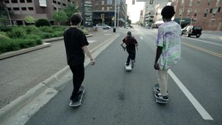 A picture of three boys skateboarding down a road surrounded by large buildings.