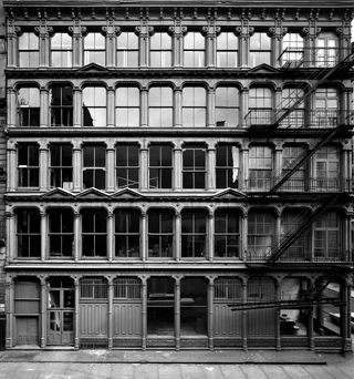A black & white building in SOHO. Large windows between each is an ornamental concrete column. There is a fire escape stairway to the right.