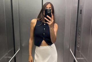 Woman standing in elevator wearing Zara black vest and cream slip skirt.