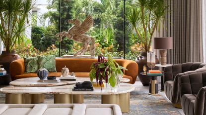 A bright living room with brown armchairs, an ochre sofa, potted dragon tree houseplants, twin coffee tables, patterned pillows, a table lamp, and a flying horse statue