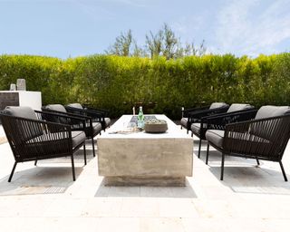 Patio space with shaped hedge in the distance, framing rectangular fire pit and black wicker chairs surrounding