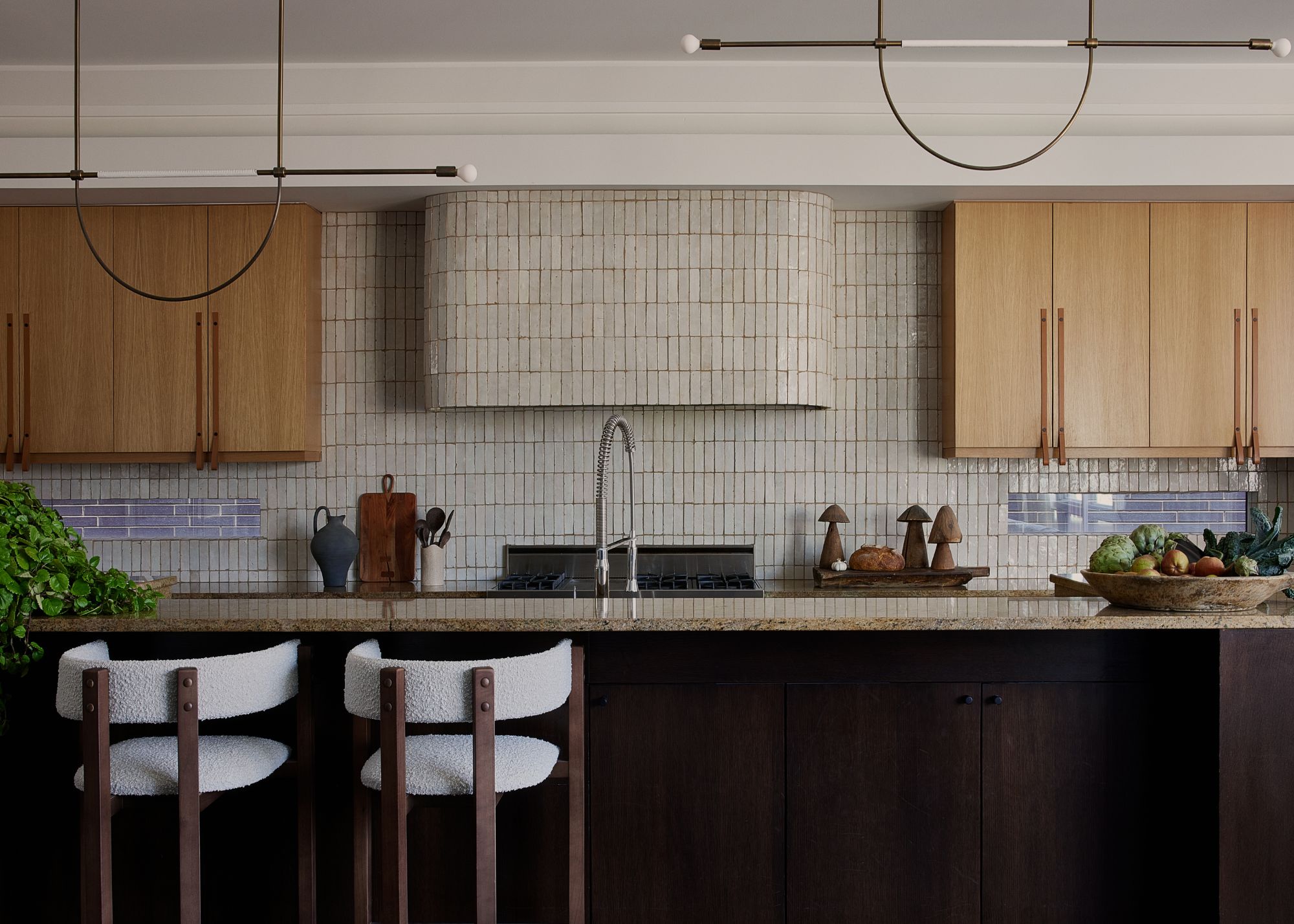 A kitchen with zellige wall tiles, wooden cabinets, granite island top and two bar stools