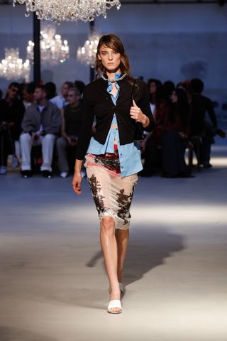 A female model wearing a black jacket, a blue button up shirt, a see through white and black lace skirt and white sandals.