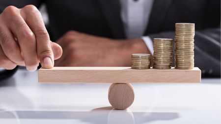 A businessman uses his finger to balance coins on one end of a small seesaw.