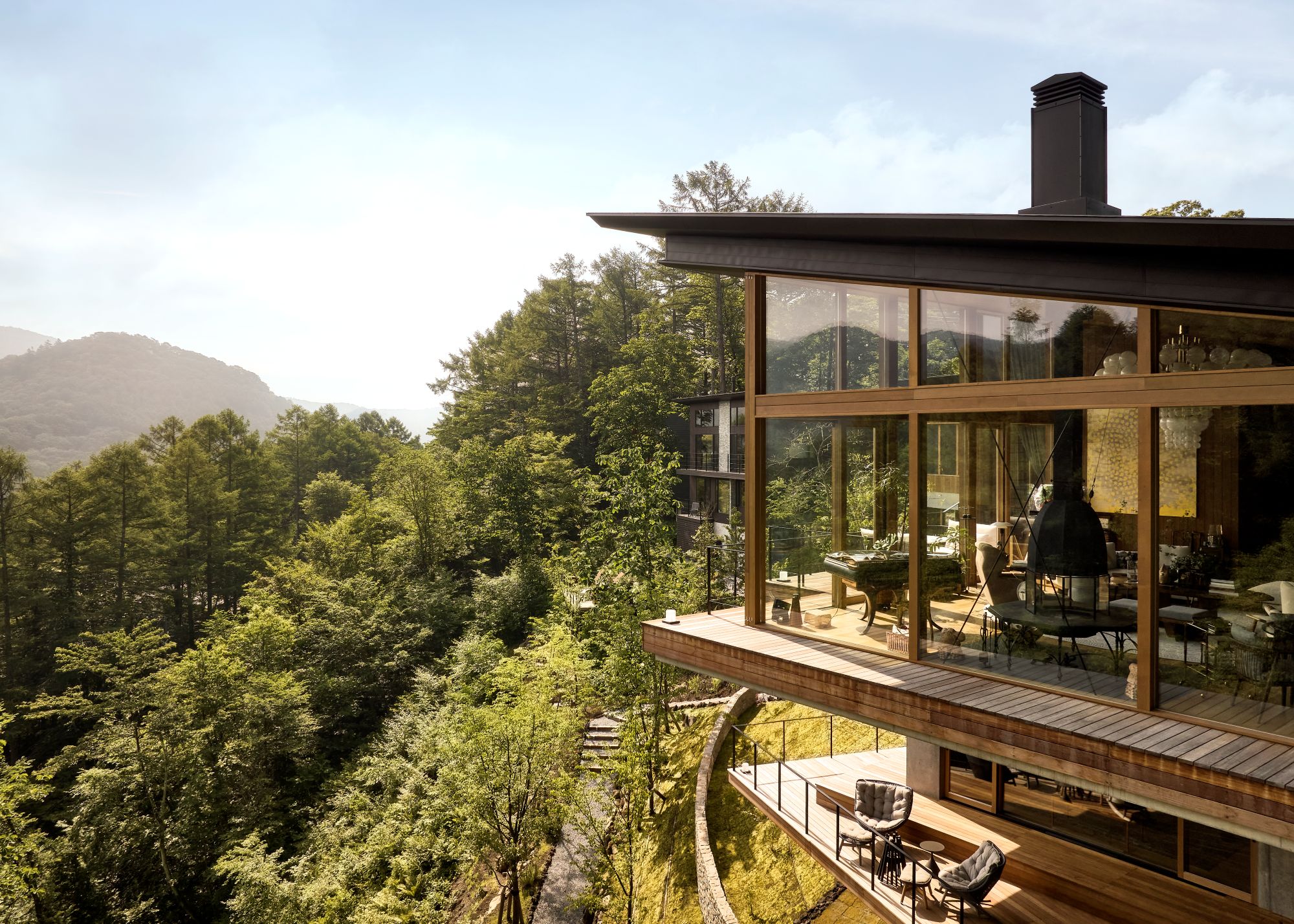 A grand Japanese home angled towards the surrounding forest and mountains with large wood-paneled glass windows all around and a sloped black roof with a black chimney breast