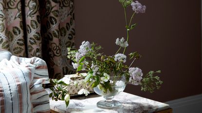 A glass vase of flowers on a marble table next to an orange, black and white striped cushion. Behind are pink, leafy patterned curtains and a terracotta wall.